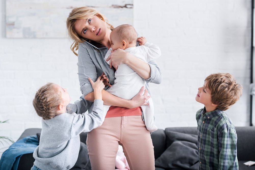 tired mother talking by smartphone and holding infant child while siblings standing in room